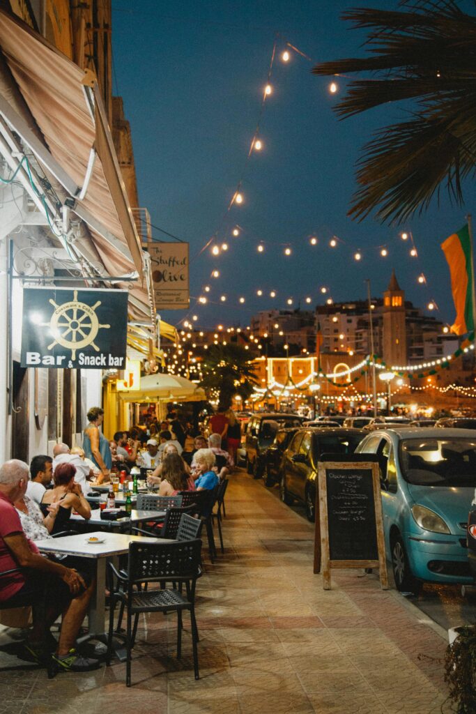 A lively street scene in Mgarr, Malta, showcasing diners enjoying an evening at a bar with string lights.